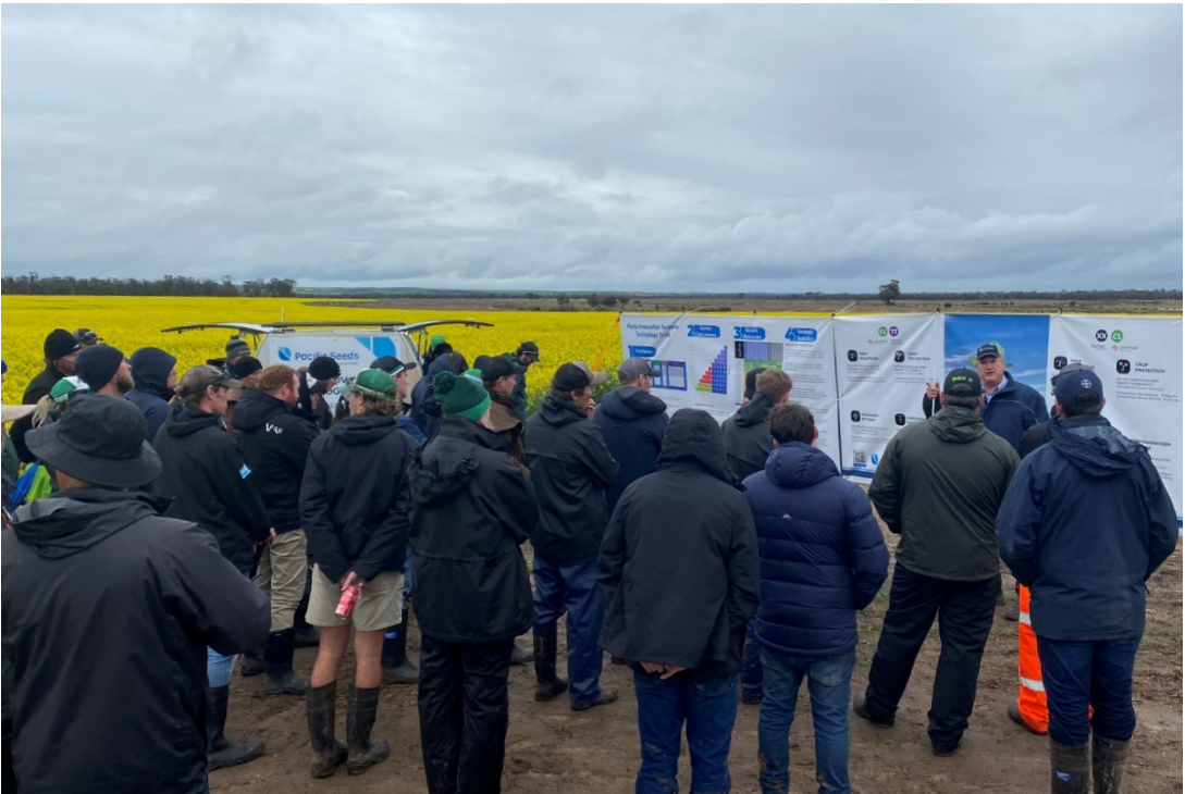 Crowd in winter coats watching an outdoor presentation beside a field under a cloudy sky