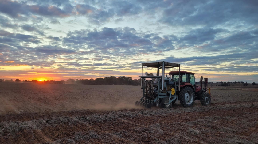 Tractor plowing a field at sunset under a cloudy sky, dust trailing behind.