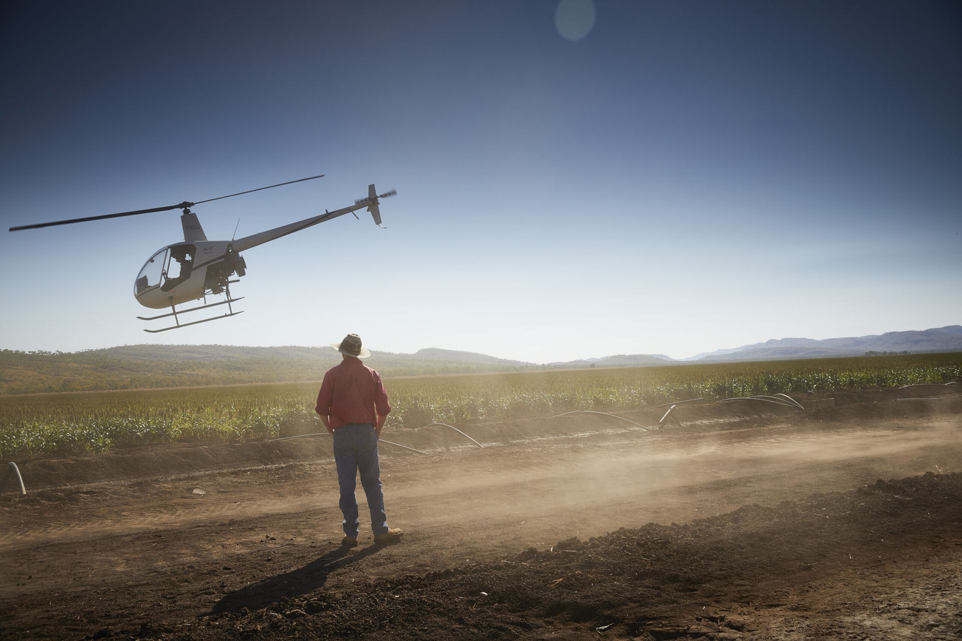 Person in red jacket watching a helicopter land on a dusty field under a clear sky.