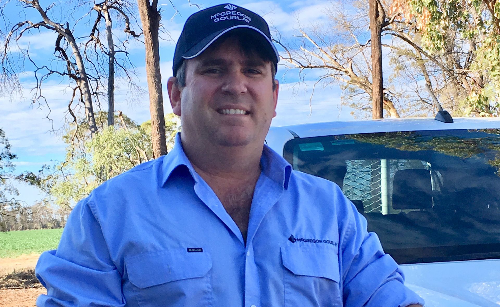 Man in blue shirt and cap standing beside a truck outdoors under a clear sky.