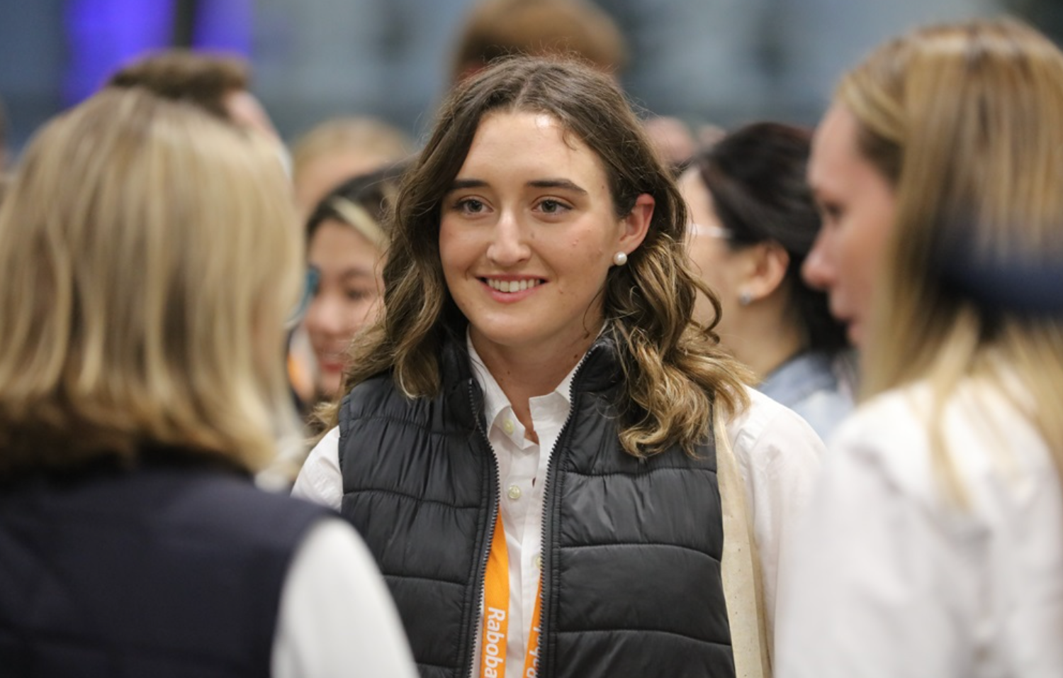 Smiling woman in a black vest stands among a small crowd indoors, with blurred people in the background