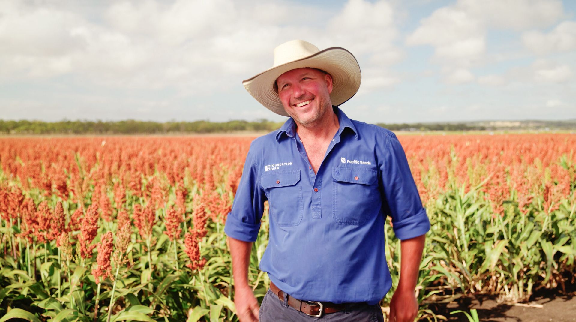 Smiling farmer in a blue shirt and cowboy hat standing in a red sorghum field under a cloudy sky