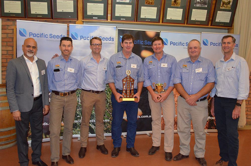 Group of people in blue shirts posing with trophies in front of a plaque wall.