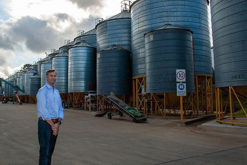 Man standing before rows of large blue industrial silos on a paved site under cloudy skies