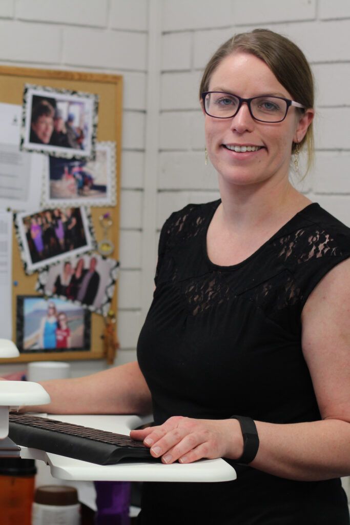 Woman in glasses smiling at a desk with a keyboard in an office, with photos pinned on a board behind her