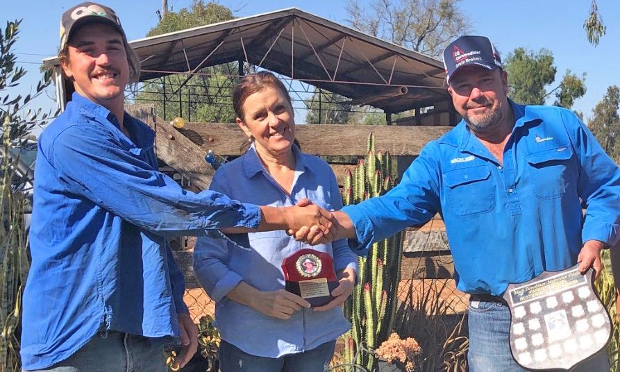 Three people in blue shirts hold a jar together outdoors in front of a greenhouse