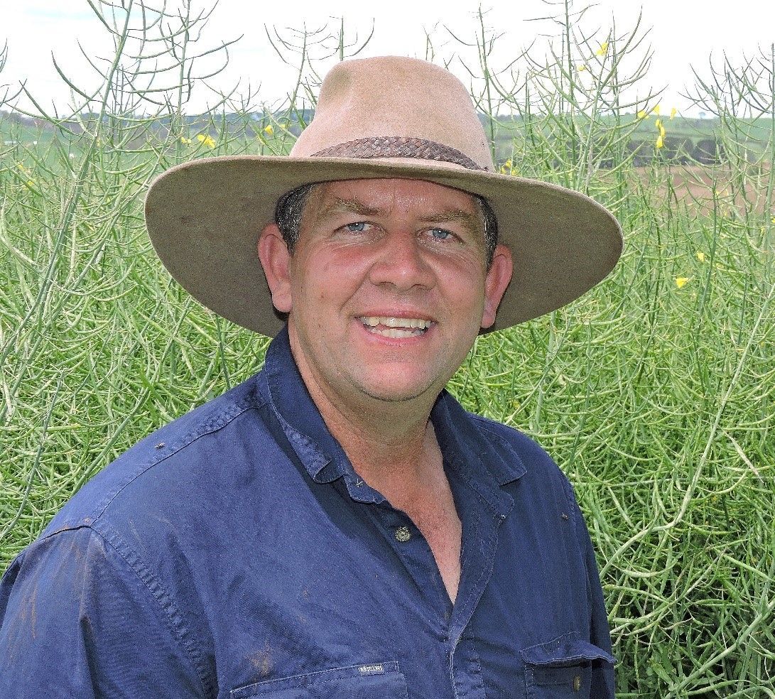 Man in a brown hat and blue shirt standing in tall green grass, smiling outdoors