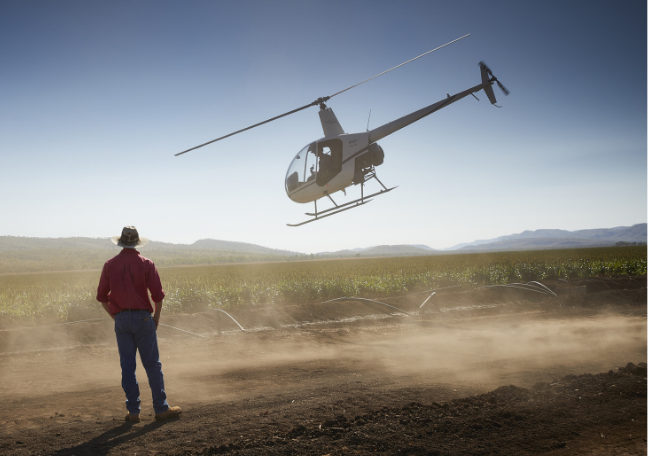 Helicopter hovering over a dusty field near a person in a red shirt under a clear sky