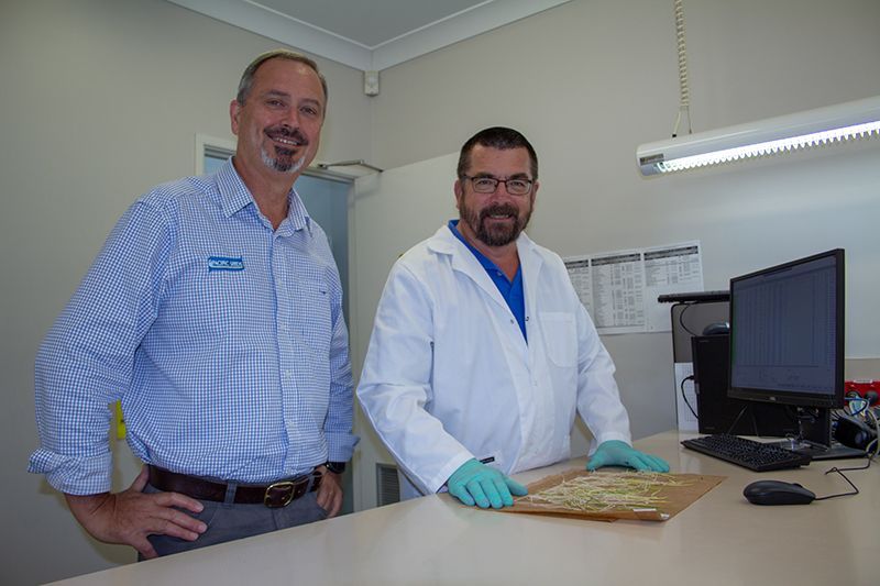 Two men in a lab, one in a white coat preparing a sample at a desk with a computer monitor.