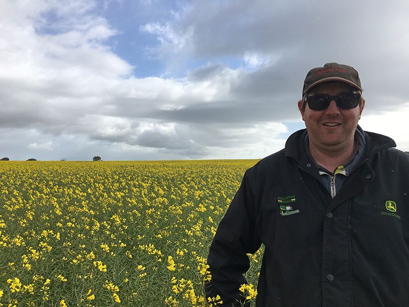 Smiling person in sunglasses and cap standing in a yellow flower field under a cloudy sky
