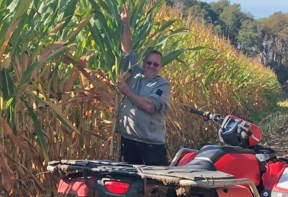 Person standing beside a red ATV in a cornfield, holding tall corn stalks.
