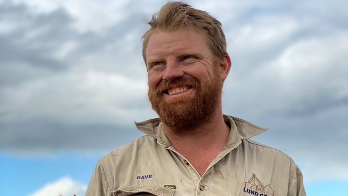 Smiling bearded man in a tan shirt standing outdoors under a cloudy sky