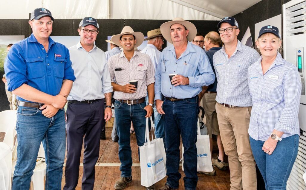 Six people in work clothes and hats stand together outdoors at a building entrance, one holding a paper bag.