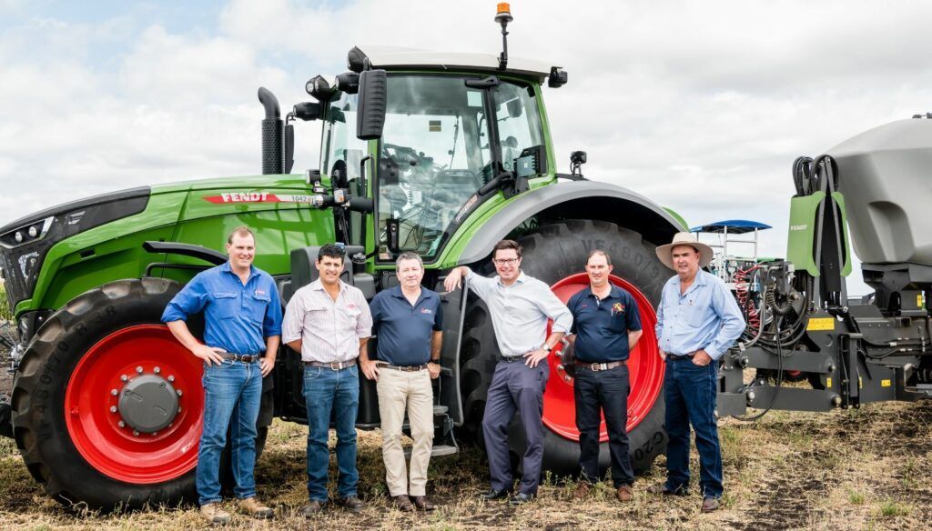 Six people posing in front of a green tractor and farm equipment in a field