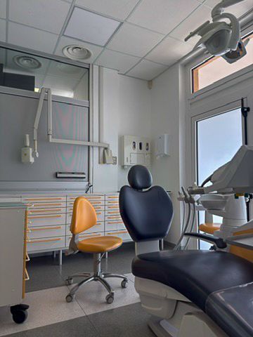 Sala visite odontoiatriche con poltrona blu, sgabello arancione e lampada a soffitto vicino a una finestra.