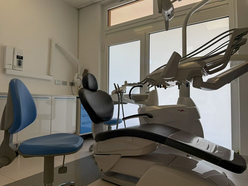 Studio dentistico con poltrona blu, sedia nera per il paziente e attrezzatura odontoiatrica vicino a una finestra.