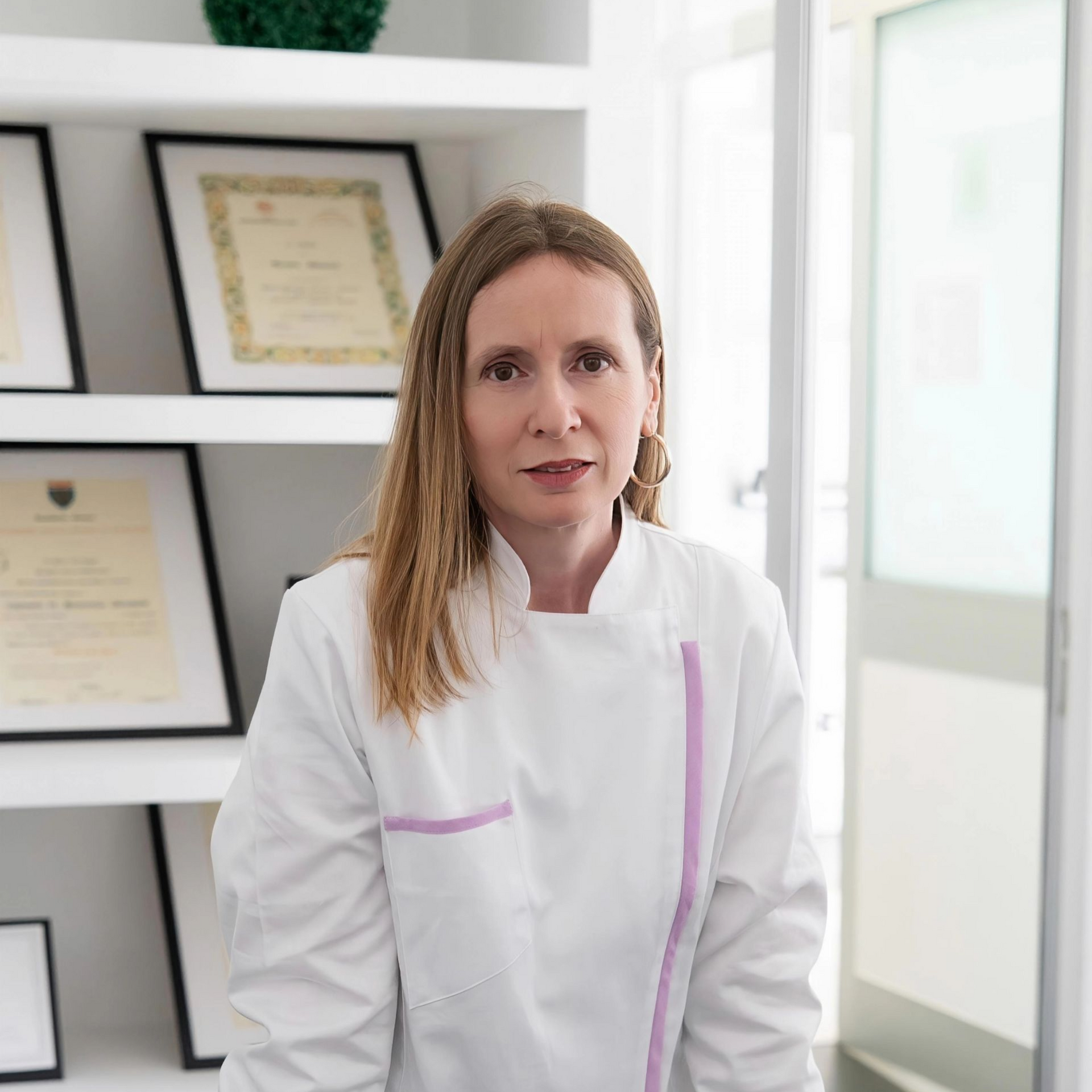 Una donna in camice bianco è in piedi in un ufficio luminoso, con certificati incorniciati appesi alla parete.