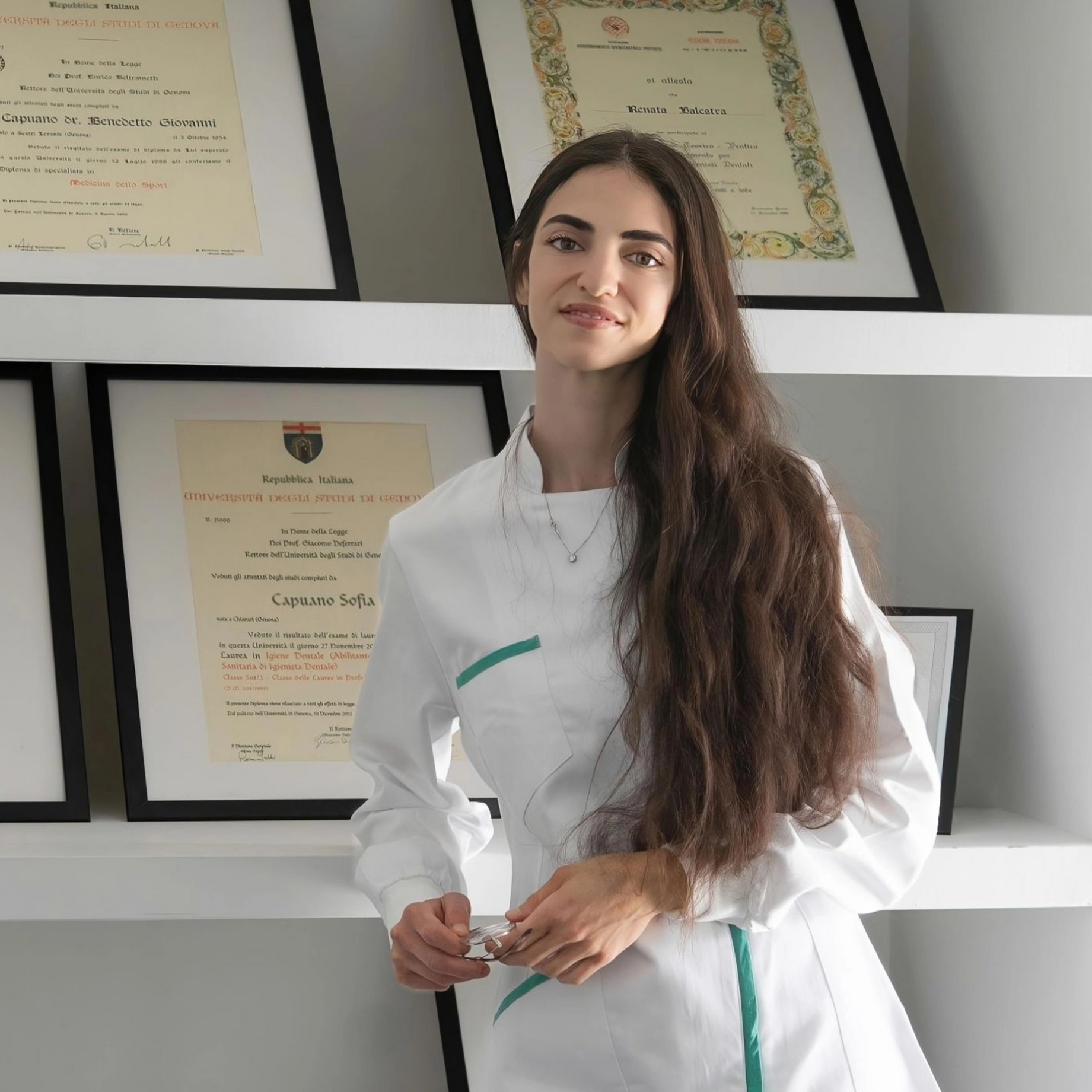 Donna in abito bianco in piedi in un ambiente interno, davanti a certificati incorniciati appesi a una parete.