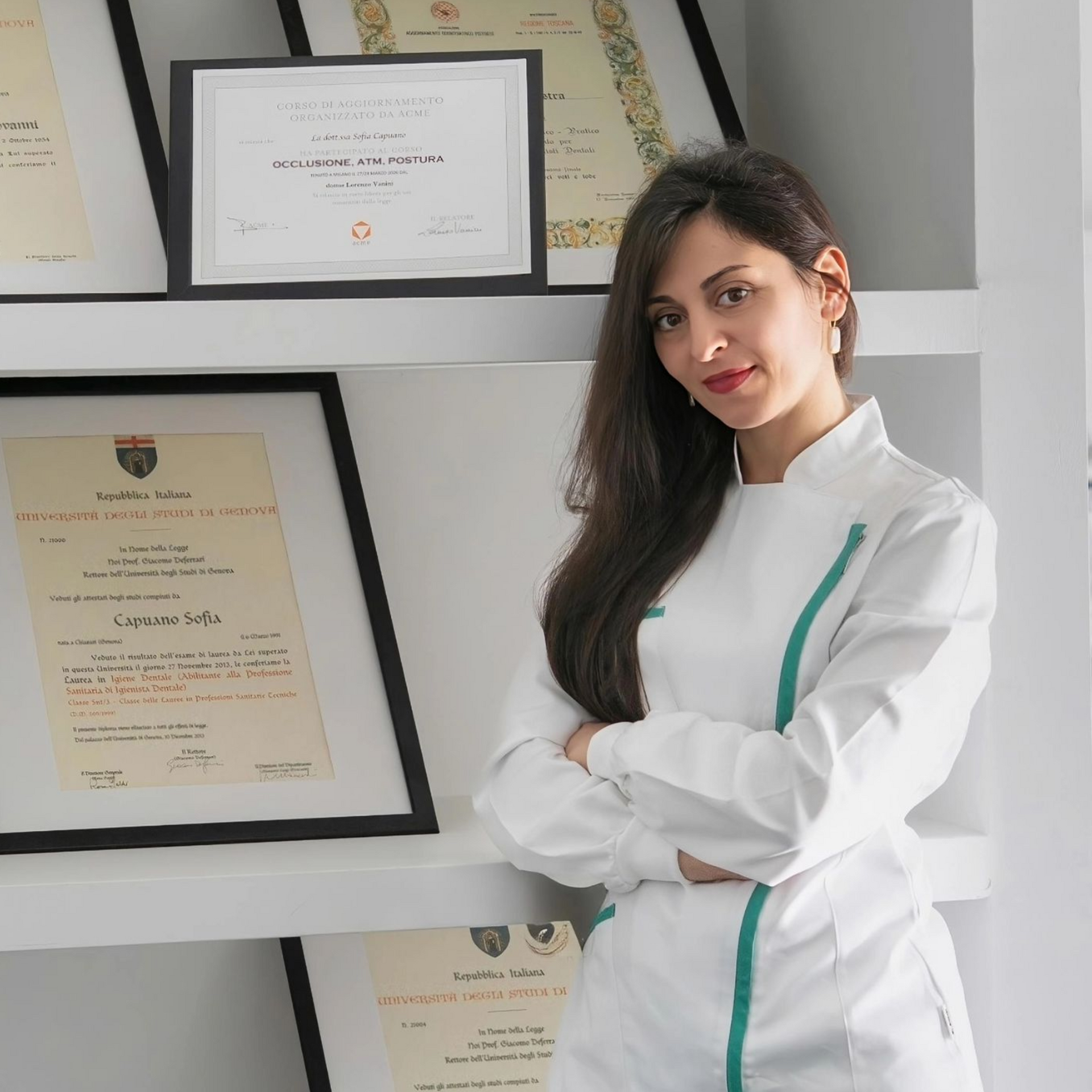 In un ufficio luminoso, una donna con una camicia bianca è in piedi, con le braccia incrociate, accanto a dei certificati incorniciati appesi a una parete.