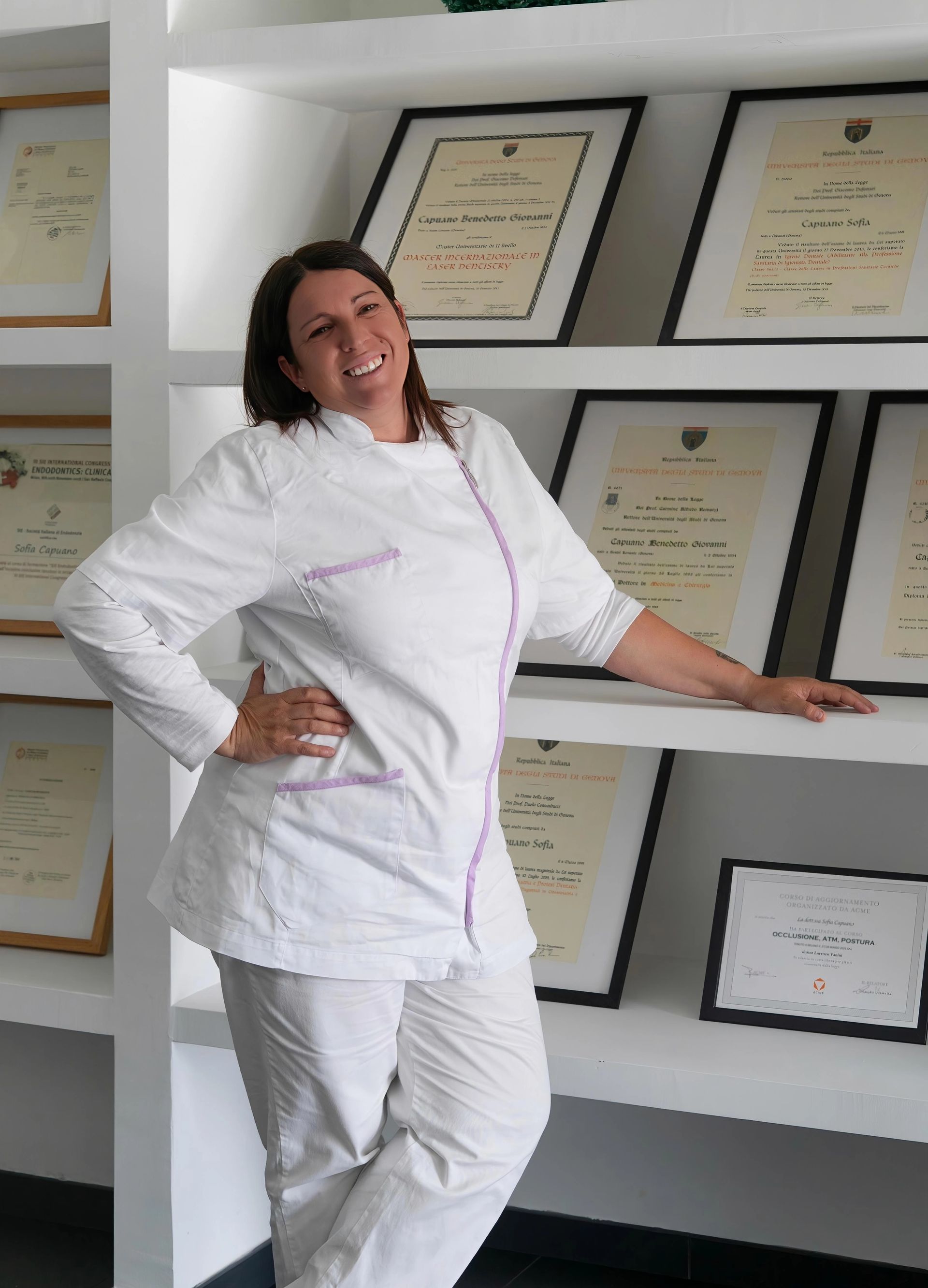 Una donna in giacca da chef bianca sorride accanto a dei certificati incorniciati appesi a una parete.
