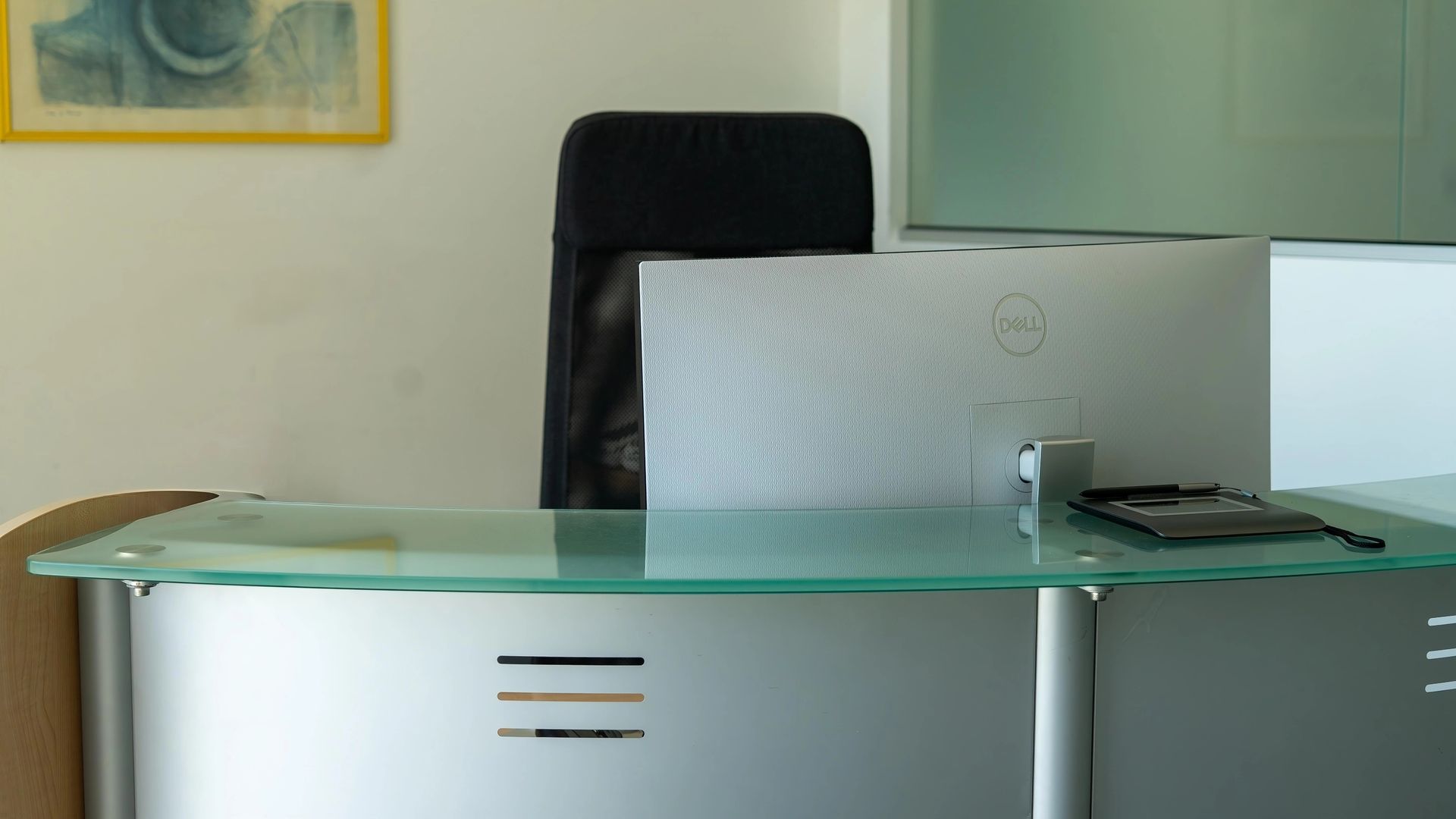 Scrivania vuota con sedia nera e monitor, contro una parete bianca.