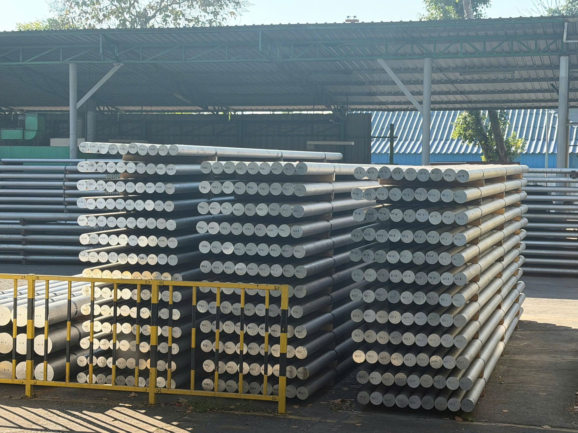 Stacks of metal pipes or poles stored outdoors under a corrugated shed, with a dirt lot and fencing behind.