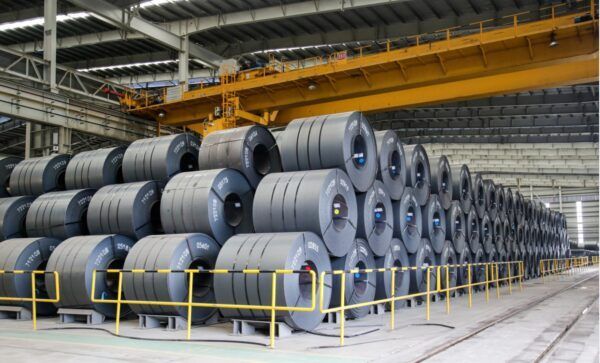 Rows of large steel coils stored in a warehouse, with a yellow overhead crane above.