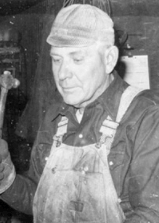 A black and white photo of a man wearing overalls and a hat holding a hammer.