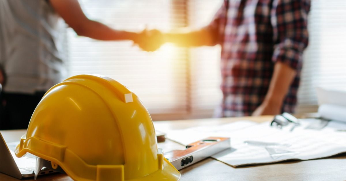 Two people shaking hands in an office. A nearby table holds a hard hat and other supplies.