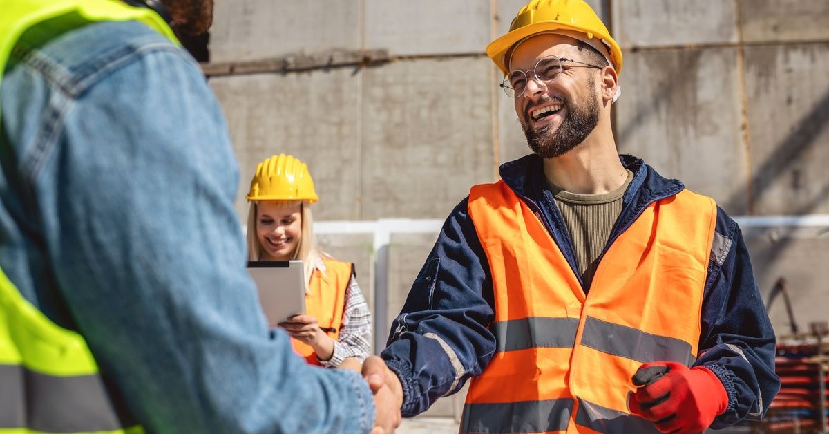 Two construction workers smiling and shaking hands on a job site. Another worker stands and smiles nearby.