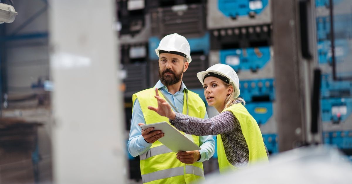 Two employees wearing hard hats standing in a brightly lit manufacturing facility. One employee hold