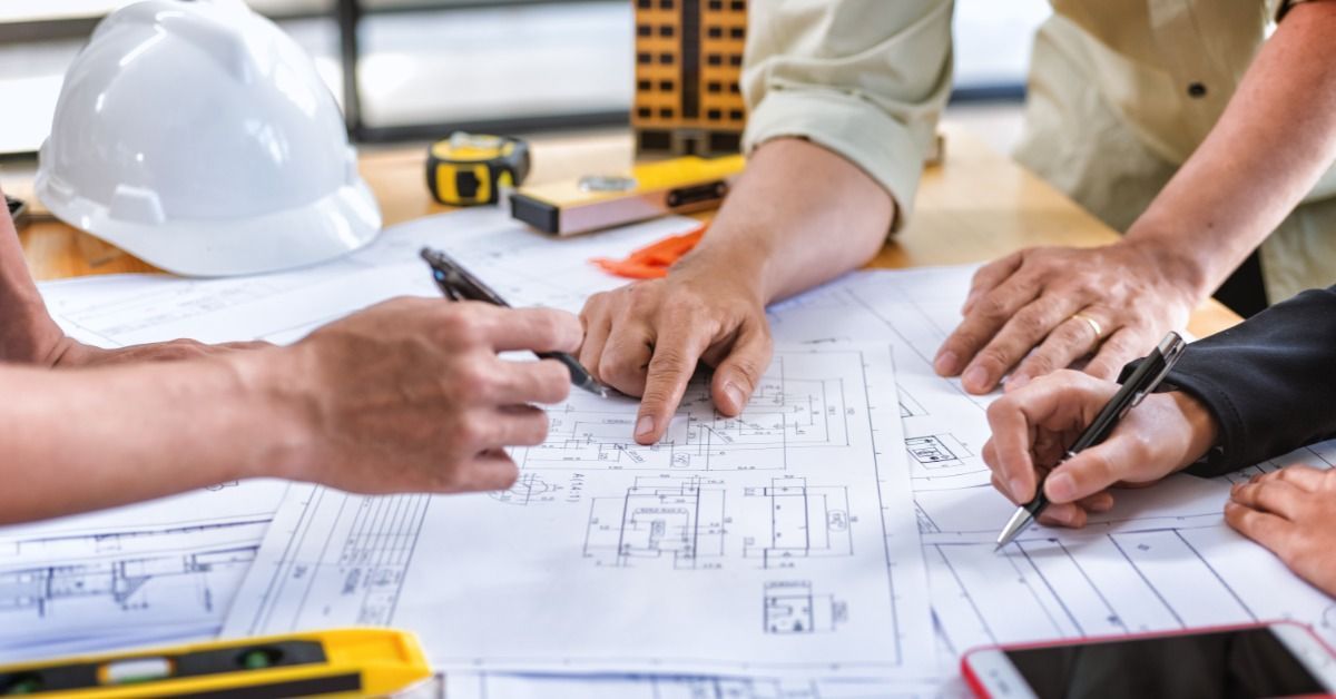 Three professionals leaning over a table covered in various blueprints. Two of the professionals have pens.