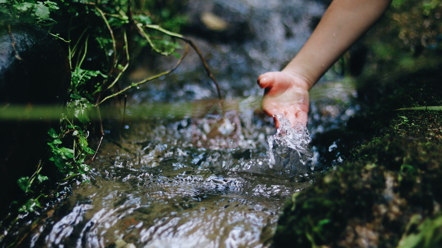 Hand reaching into a clear stream with water splashing over rocks and green moss