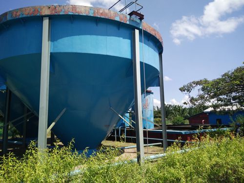 Blue industrial storage tank under a partly cloudy sky, surrounded by grass and trees
