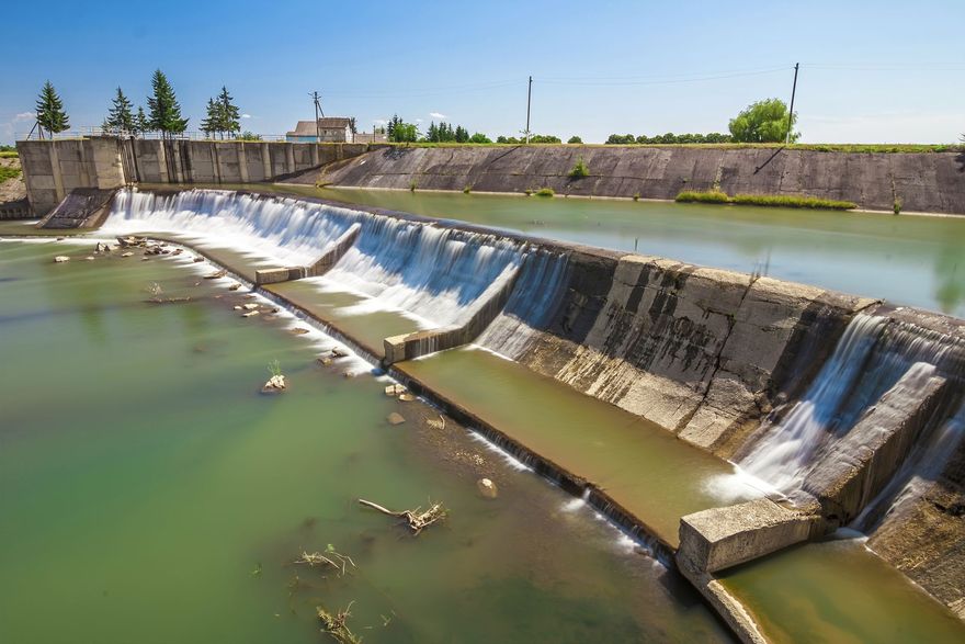 Low dam spilling water into a green canal under a blue sky