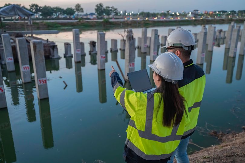 Two workers in hard hats and high-vis jackets inspect a waterfront piling area at dusk.