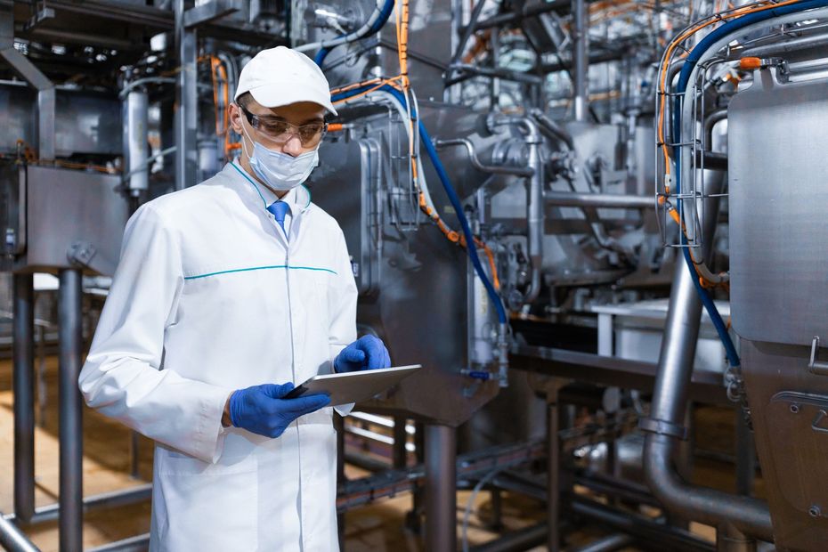 Technician in white lab coat and hard hat checks equipment in an industrial plant.