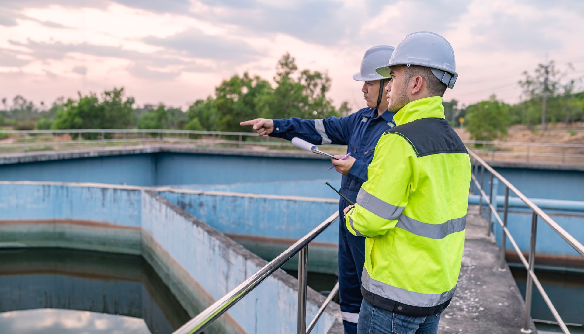 Two workers in hard hats inspect a water treatment tank, one pointing across the basin.