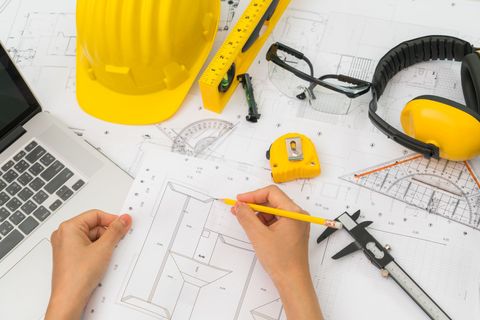 Hands sketching architectural plans with yellow hard hat, ruler, laptop, and safety glasses on a desk