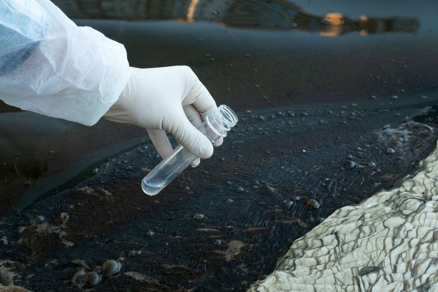 Gloved hand holding a sample vial over a dark polluted water shoreline