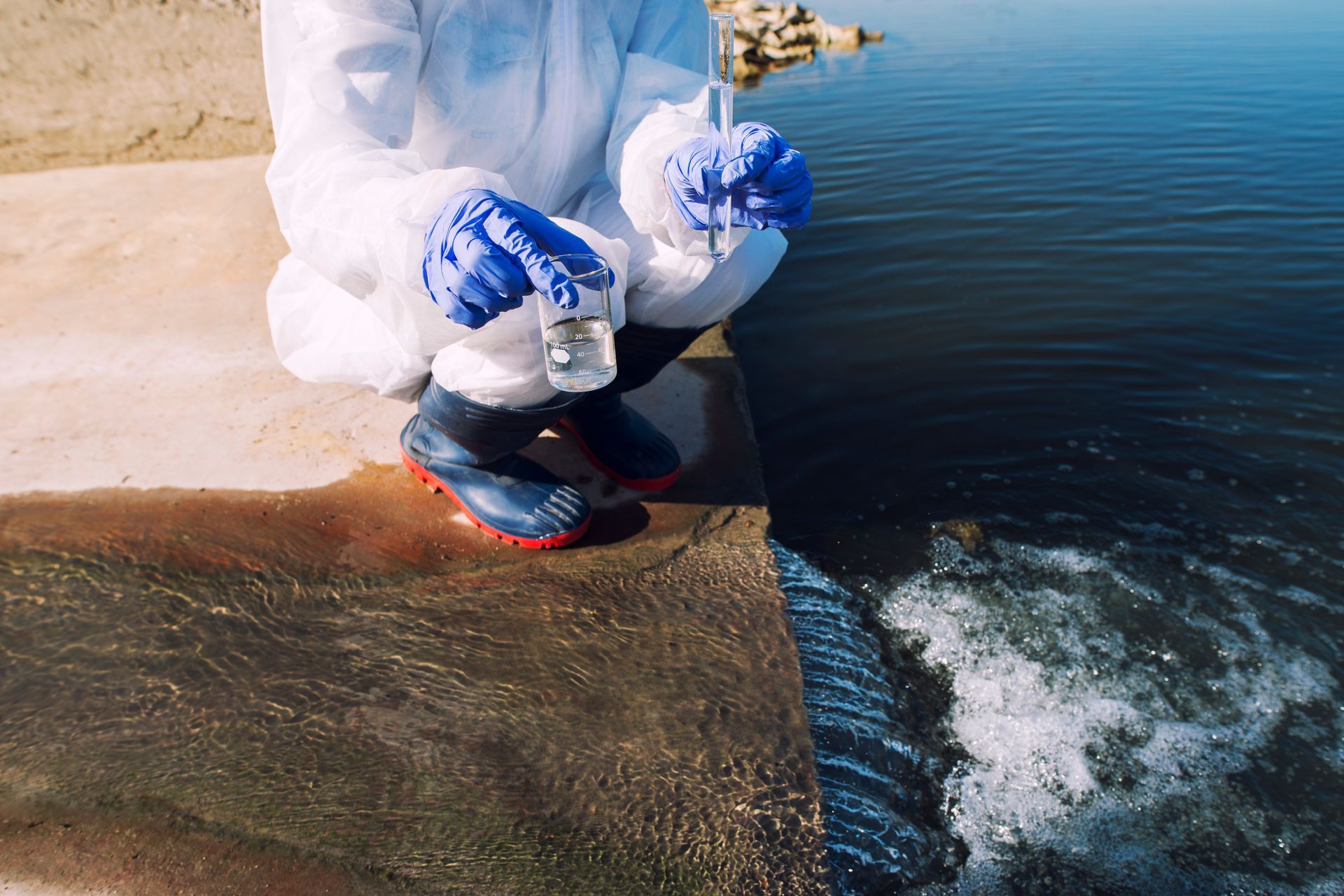 Person in white protective suit crouches by a concrete edge above dark blue water, holding a blue net.