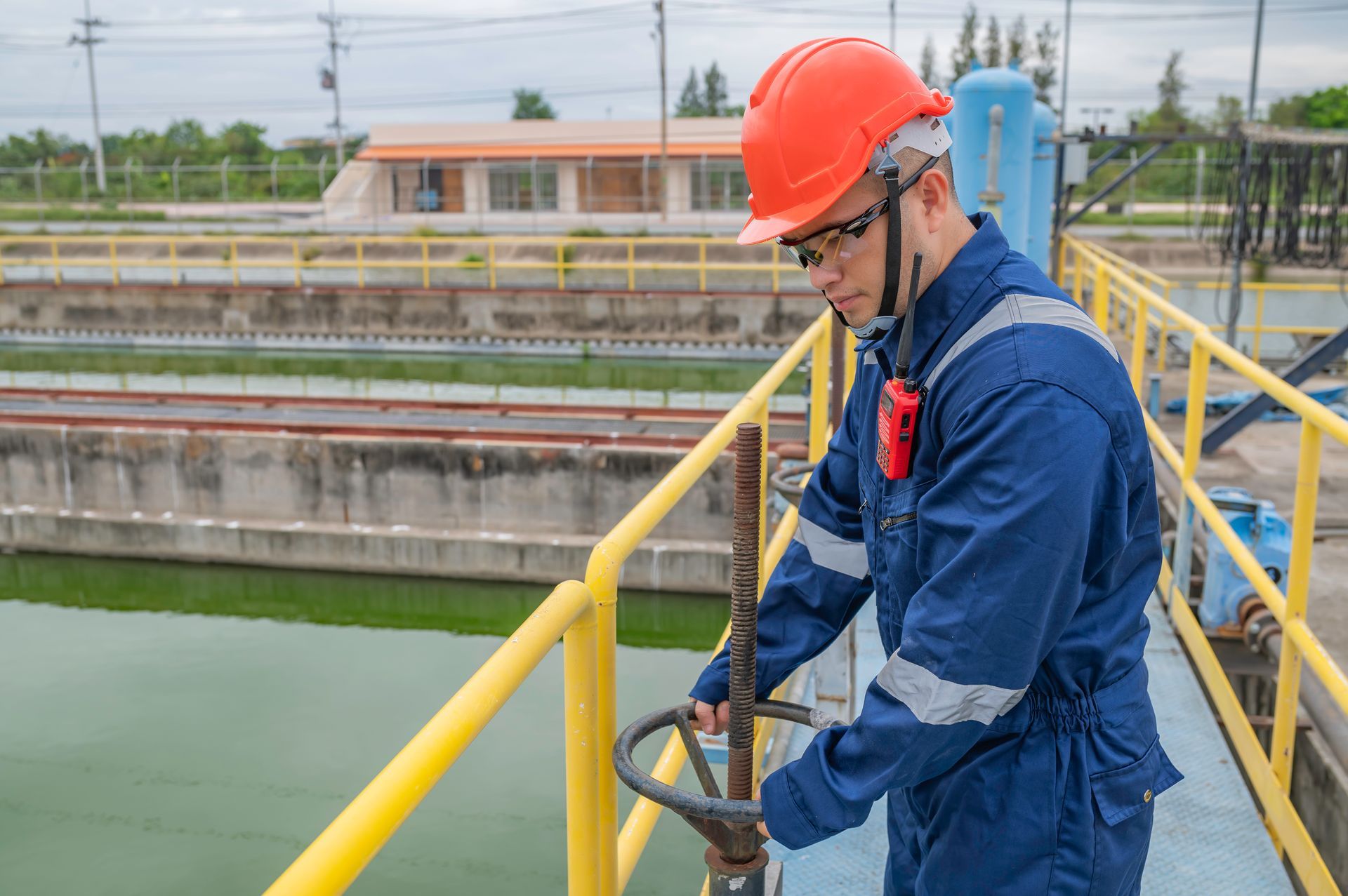 Worker in blue safety gear and orange helmet inspecting equipment at an outdoor treatment facility.