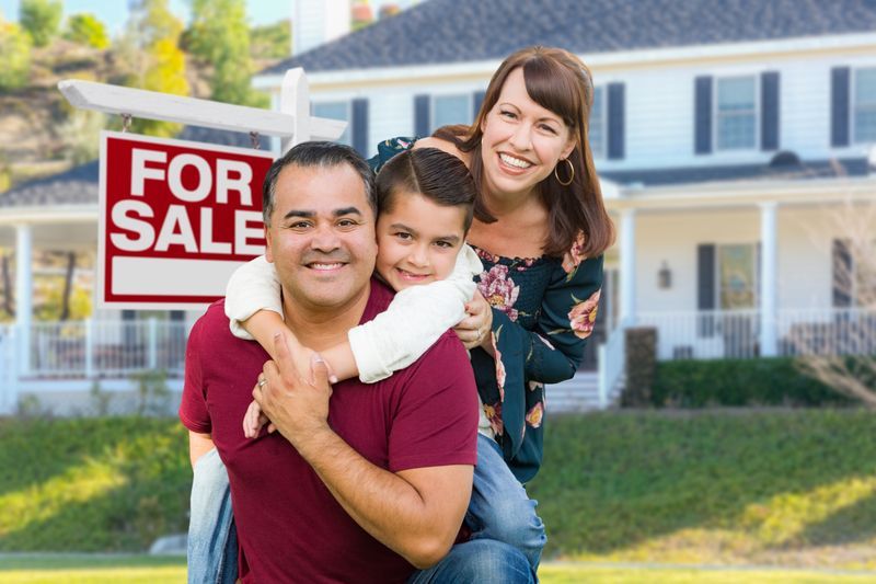 A man is carrying a child on his back in front of a for sale sign.