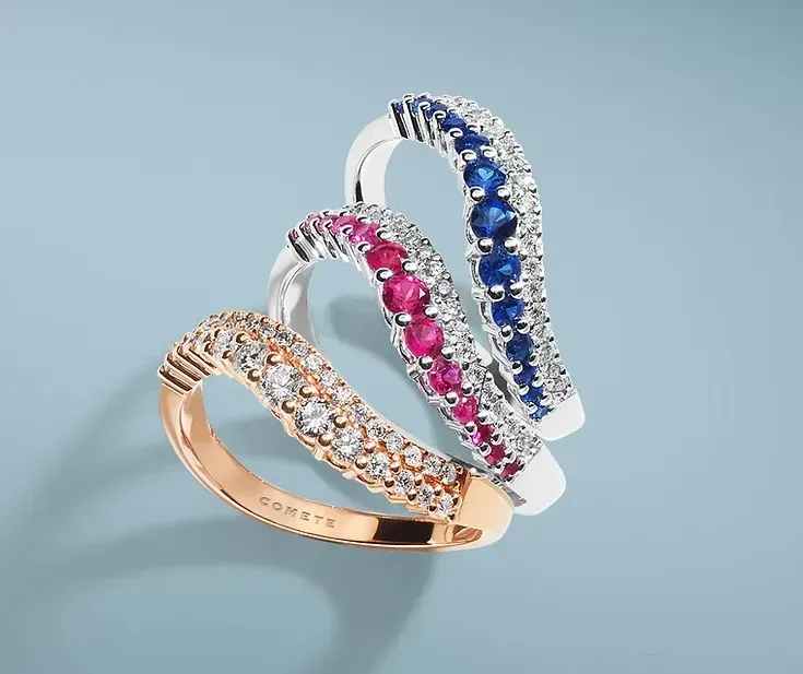 Tre anelli con gemme rosa, blu e trasparenti su uno sfondo azzurro chiaro