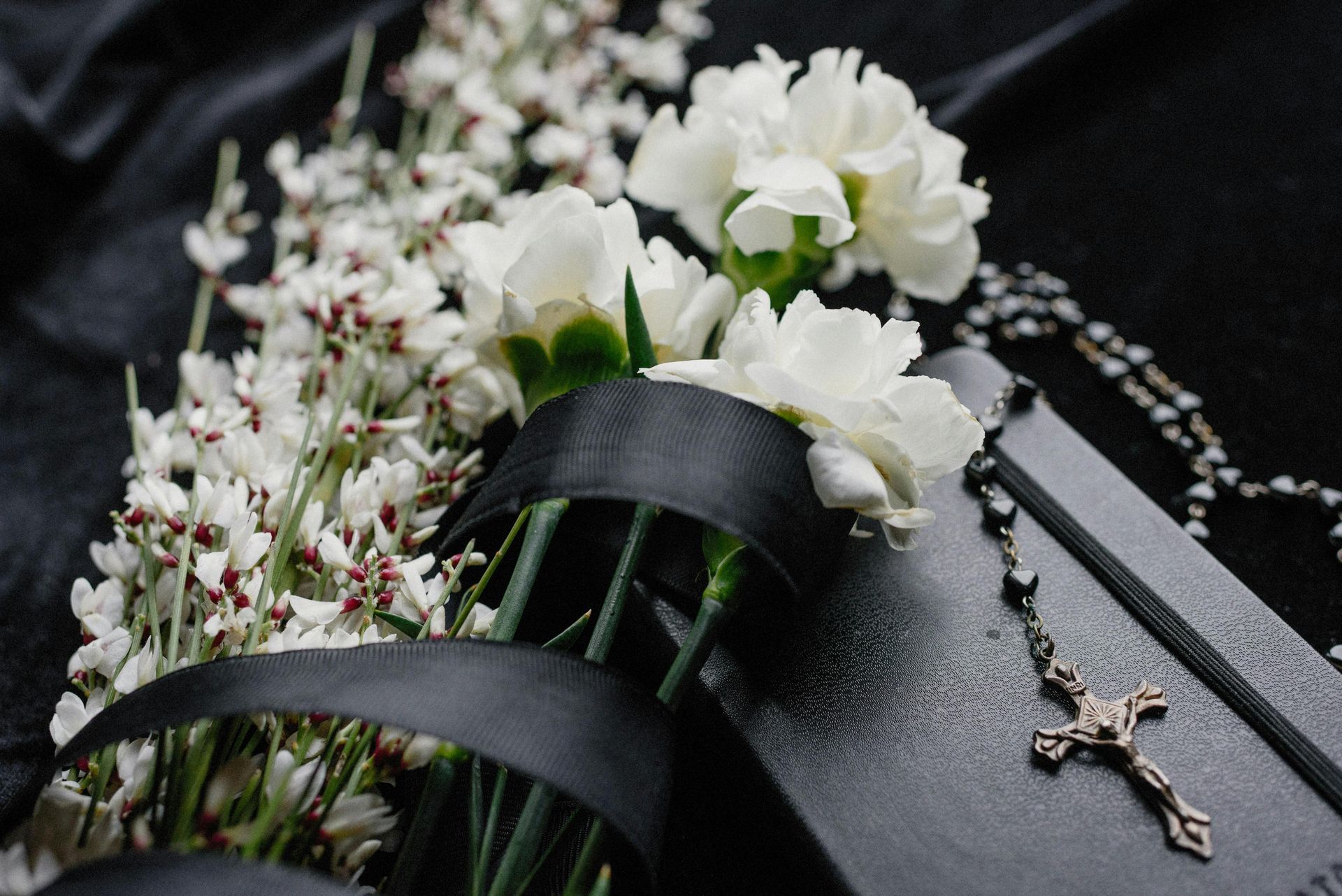 White flowers and a black ribbon on a dark surface, with a rosary and a book.