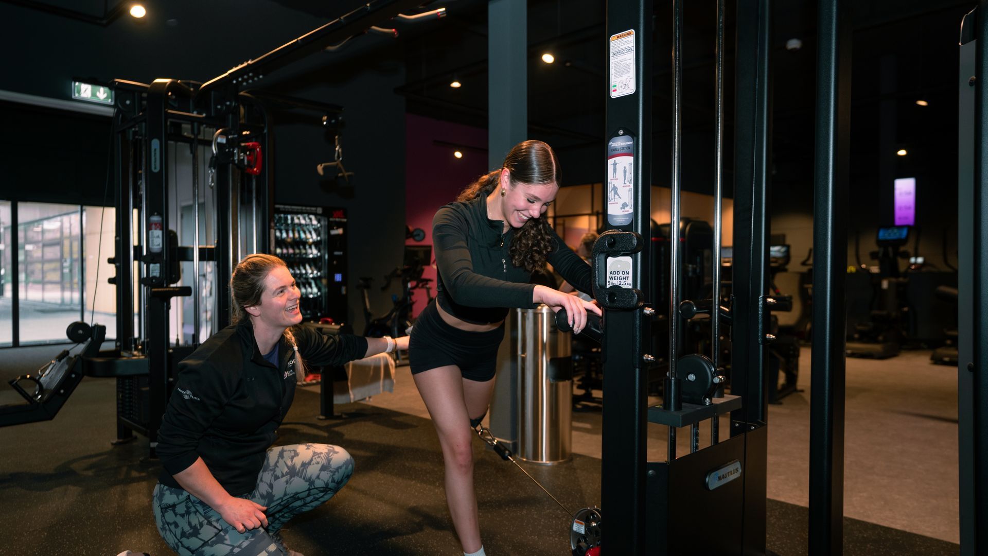 Woman using cable machine in gym, supervised by another woman.