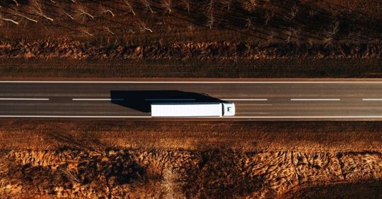White semi-truck driving on a straight highway through a dry, brown landscape viewed from above