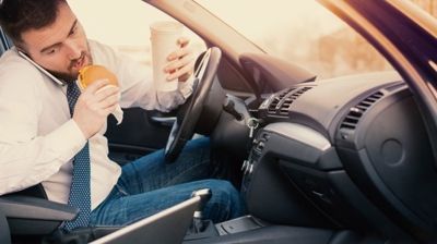 Driver eating a burger and holding a drink while seated in a car