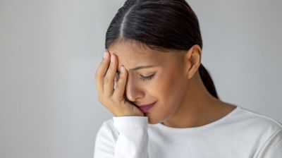 Woman in a white shirt holding her forehead with eyes closed, looking stressed against a plain background