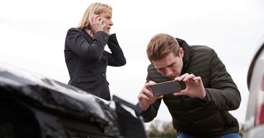 Two people outdoors, one photographing a car bumper while the other looks on in concern